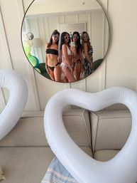 Mirror selfie of four women in bikinis and heart-shaped sunglasses, one wearing a veil, posing in a bright coastal-style hotel suite with colorful balloons and a large white inflatable heart — bachelorette party vibe.