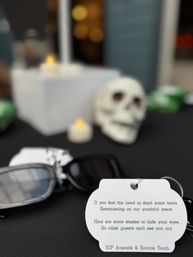 Close-up of a party favor table with sunglasses and a printed tag offering 'shades' for tears, LED candles and a decorative skull — playful memorial/Halloween display.