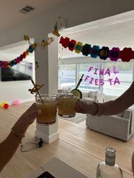 Two hands clinking salt-rimmed cocktails with lime and paper straws in a sunlit living room decorated with colorful paper-flower garlands and 'FINAL FIESTA' window balloons, party balloons and a bottle on the table.