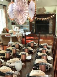 Cozy living room party setup: wooden dining table arranged with rose-gold insulated tumblers and drawstring favor bags, decorative paper fans and bunting hanging near a fireplace.