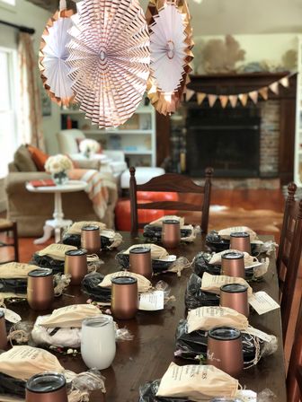 Cozy living room party setup: wooden dining table arranged with rose-gold insulated tumblers and drawstring favor bags, decorative paper fans and bunting hanging near a fireplace.