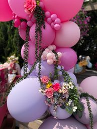 Pink and lavender balloon garland with cascading green vines and a mixed floral cluster of roses and wildflowers, styled for an outdoor party or garden celebration