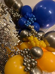 Close-up of a festive balloon garland with large royal blue and bright yellow balloons, metallic silver cluster accents, and a sparkling mirrored sequin backdrop for event party decor.