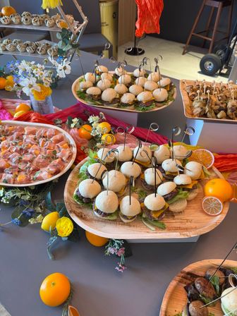 Colorful catered buffet table with wooden platters of mini cheeseburger sliders on skewers, prosciutto-wrapped bites, grilled skewers, fresh oranges and floral decor — stylish appetizer spread for an event.