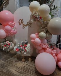 Indoor baby shower backdrop with pastel pink and beige balloon garland, floral accents, wicker baby pram and decorative Oh Baby sign