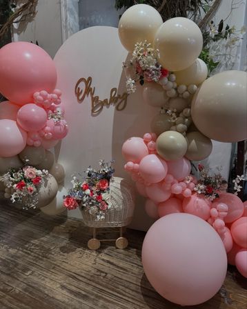Indoor baby shower backdrop with pastel pink and beige balloon garland, floral accents, wicker baby pram and decorative Oh Baby sign