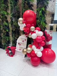 Valentine's Day indoor photo backdrop with large red, white and gold balloon garland, lush greenery wall, white display shelf with champagne flutes and red-draped table.