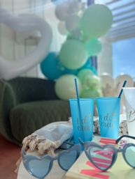 Indoor bachelorette party scene with two blue "I do crew" cups and straws, heart-shaped sunglasses, a textured throw, and teal-and-white balloon garland in soft focus
