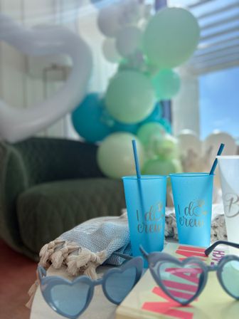 Indoor bachelorette party scene with two blue "I do crew" cups and straws, heart-shaped sunglasses, a textured throw, and teal-and-white balloon garland in soft focus