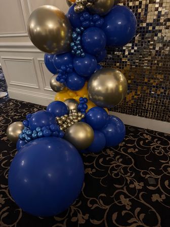 Cascading navy blue, metallic gold and yellow balloon garland event display against a sequin backdrop on patterned black ballroom carpet