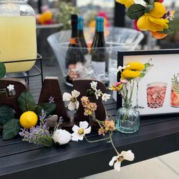 Rooftop bar display on an outdoor table with wooden 'BAR' letters, lemons and mixed flowers, glass vase of yellow blooms, ice bucket with wine bottles and a lemonade dispenser — stylish cocktail setup.