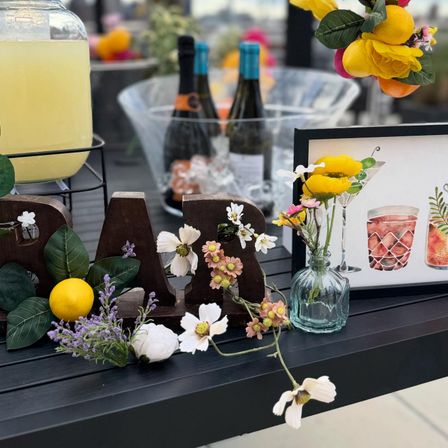 Rooftop bar display on an outdoor table with wooden 'BAR' letters, lemons and mixed flowers, glass vase of yellow blooms, ice bucket with wine bottles and a lemonade dispenser — stylish cocktail setup.