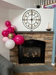 Cheerful pink, white and gold balloon cluster on a stone fireplace mantel in a cozy living room, topped by a large metal wall clock and a small sailboat model.