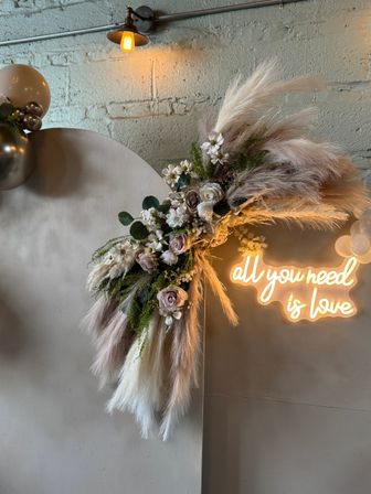 Boho wedding/event backdrop with pampas grass and neutral roses, metallic balloons, warm Edison bulb, and neon 'all you need is love' sign against an industrial brick wall