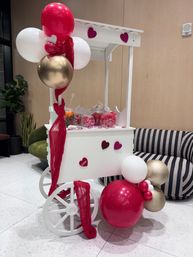 Valentine's-themed white candy cart in a modern indoor lobby, decorated with red, pink, white and gold balloons, heart accents, draped red fabric and glass jars of pink candy beside a striped sofa