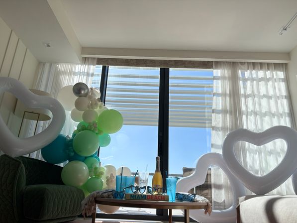 Sunlit coastal living room decorated for a party with green-and-white balloon garland, white heart-shaped inflatables, sheer curtains framing sliding glass doors to blue sky and ocean, and a small table with champagne and blue cups.