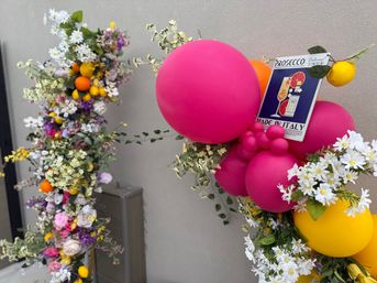 Bright balloon and floral arch with large pink and yellow balloons, white daisies, faux lemons and a Prosecco sign on a neutral wall, event storefront decor