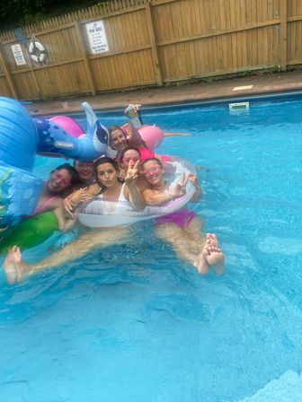 Group of friends relaxing in a backyard pool on colorful inflatable floats (unicorn and donut), smiling and flashing peace signs by a wooden fence.