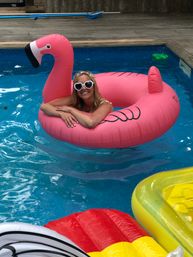 Cheerful person lounging on a giant pink flamingo float in a backyard pool, wearing heart-shaped sunglasses and a floral headband with colorful inflatable rafts nearby.