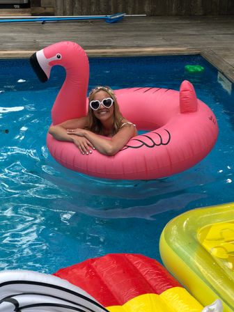Cheerful person lounging on a giant pink flamingo float in a backyard pool, wearing heart-shaped sunglasses and a floral headband with colorful inflatable rafts nearby.