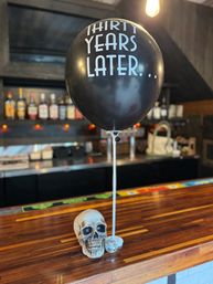 Black balloon printed 'THIRTY YEARS LATER...' tied to a stand above a decorative skull and tiny disco ball on a polished wood bar counter, with blurred liquor bottles and warm lighting in the background.