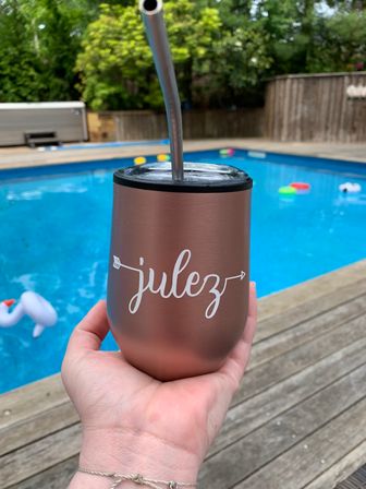 Rose-gold insulated tumbler with metal straw and white script held over a backyard swimming pool, colorful floats and wooden deck in the background