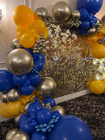 Vibrant indoor balloon arch of royal blue, yellow and metallic silver balloons framing a shimmering gold sequin photo wall — festive event backdrop.