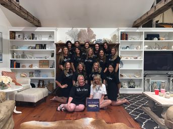 Smiling group photo of women in matching shirts gathered in a coastal beach‑house living room with white built‑in shelves of shells and nautical decor, exposed wooden beams, hardwood floors and a patterned rug.