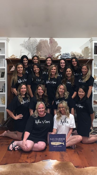 Smiling group photo of women in matching shirts gathered in a coastal beach‑house living room with white built‑in shelves of shells and nautical decor, exposed wooden beams, hardwood floors and a patterned rug.