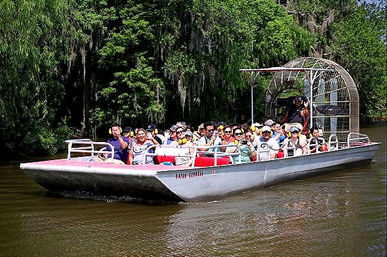 Airboat packed with tourists wearing ear protection on a sunny Louisiana bayou swamp tour, cruising past moss-draped cypress and dense green foliage.