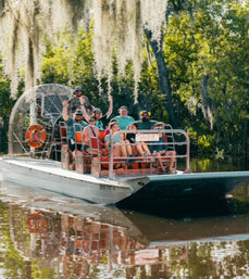 Group waving on an airboat tour gliding through a Spanish moss-draped Southern bayou, with calm water reflecting the boat and trees.