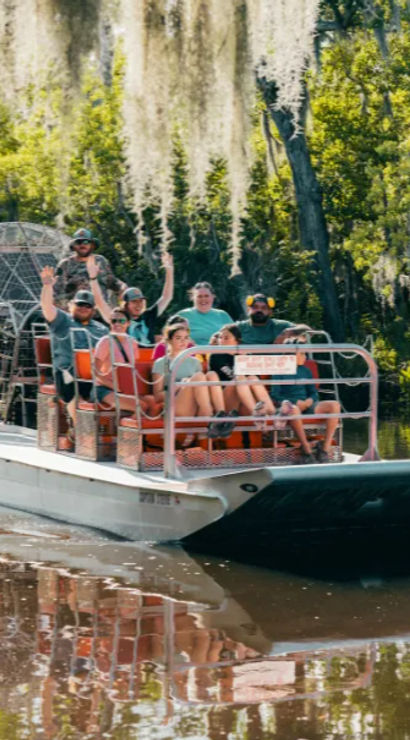 Group waving on an airboat tour gliding through a Spanish moss-draped Southern bayou, with calm water reflecting the boat and trees.