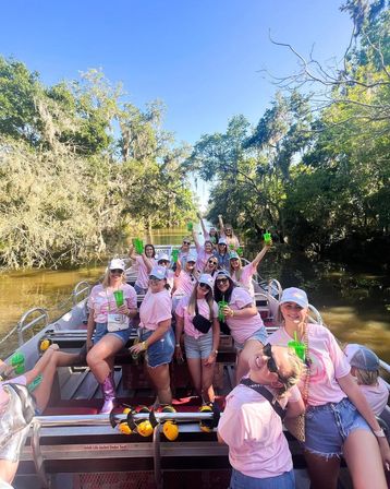 Group of women in matching pink shirts and caps enjoying a festive boat party on a sunny cypress-lined river with Spanish moss-draped trees in a southern swamp setting.