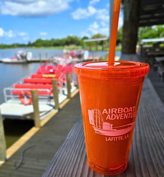 Bright orange souvenir cup with straw on a wooden dock in the foreground, blurred red airboats moored on a calm Louisiana bayou and sunny blue sky in the background.