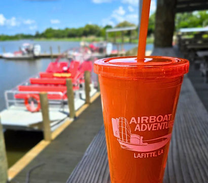 Bright orange souvenir cup with straw on a wooden dock in the foreground, blurred red airboats moored on a calm Louisiana bayou and sunny blue sky in the background.