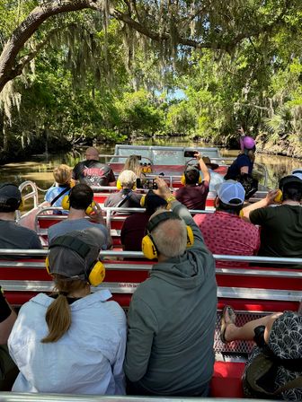 Group of people on a red airboat wearing yellow ear protection, cruising a moss-draped Florida cypress swamp beneath sunlit live oaks