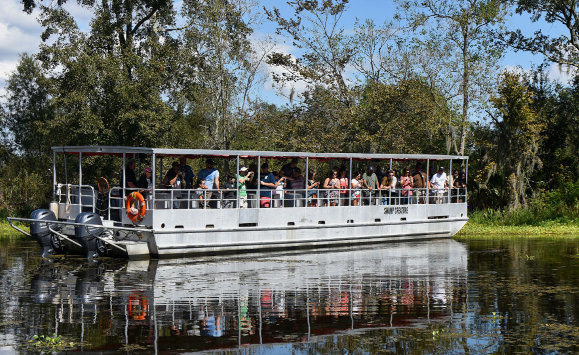 Crowded swamp tour boat cruising a tree-lined Florida marsh, passengers watching wildlife with calm water reflecting the boat and blue sky