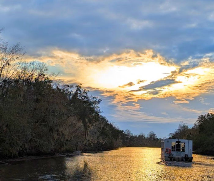 Golden sunset over a calm river framed by moss-draped cypress trees, a small houseboat drifting toward the glowing horizon.