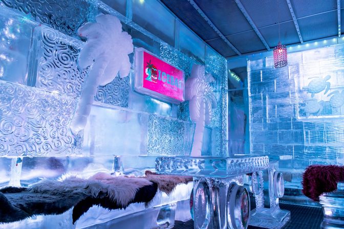 Icy indoor ice bar lounge with carved ice palm trees, sculpted ice table and benches draped in faux fur, glowing pink sign and cool blue ambient lighting.
