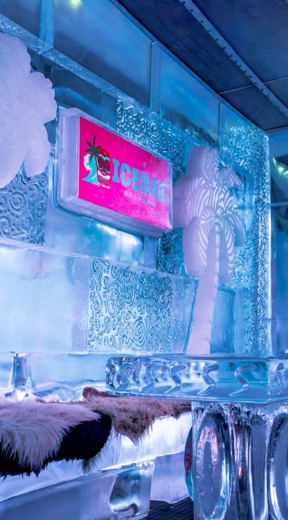 Icy indoor ice bar lounge with carved ice palm trees, sculpted ice table and benches draped in faux fur, glowing pink sign and cool blue ambient lighting.