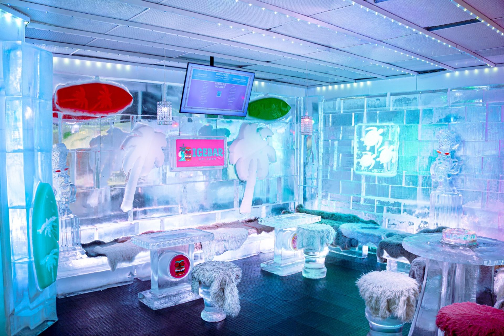 Icy cocktail lounge with carved ice walls, fur-covered stools, ice tables, surfboard and palm sculptures, and colorful blue‑pink neon lighting.