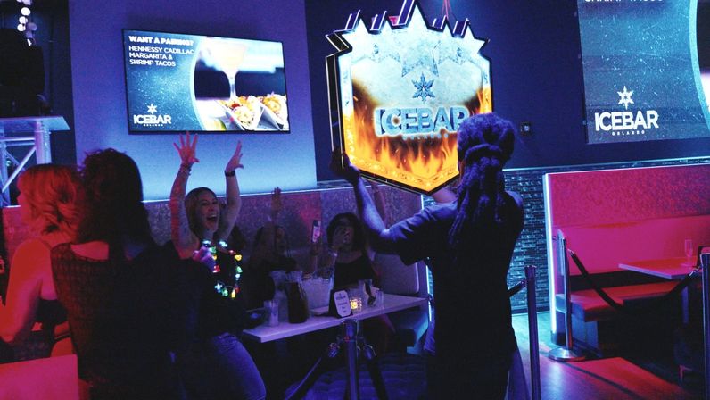 Group of people cheering as a server carries a glowing ice-themed sign through a neon-lit bar in Orlando, blue-purple lighting and drinks on the table — lively nightlife scene.