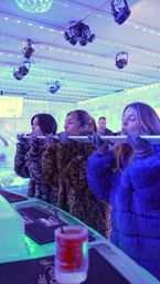 Three women in faux-fur coats and gloves taking shots from a long ice 'shot ski' at a blue-lit indoor ice bar with string LED lights and overhead stage lighting, a frozen cocktail in the foreground.