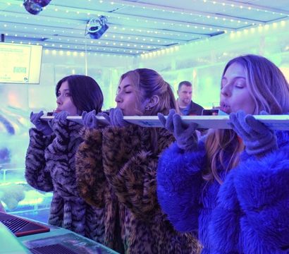 Three women in faux-fur coats and gloves taking shots from a long ice 'shot ski' at a blue-lit indoor ice bar with string LED lights and overhead stage lighting, a frozen cocktail in the foreground.