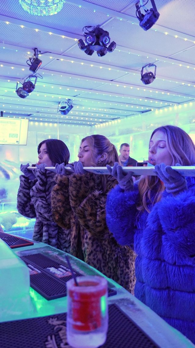 Three women in faux-fur coats and gloves taking shots from a long ice 'shot ski' at a blue-lit indoor ice bar with string LED lights and overhead stage lighting, a frozen cocktail in the foreground.