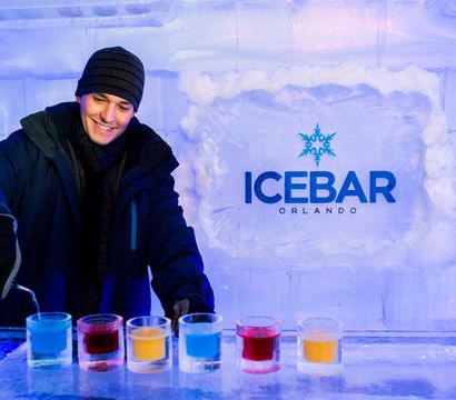 Smiling person in a beanie and gloves pouring colorful frozen cocktails at an indoor ice bar in Orlando with carved ice walls and illuminated ice blocks