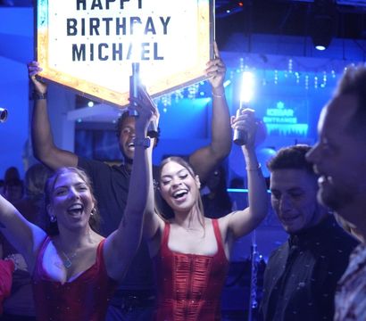 Energetic nightclub birthday celebration with smiling partygoers holding an illuminated birthday sign, bottle service and handheld sparklers under blue club lighting and balloons.