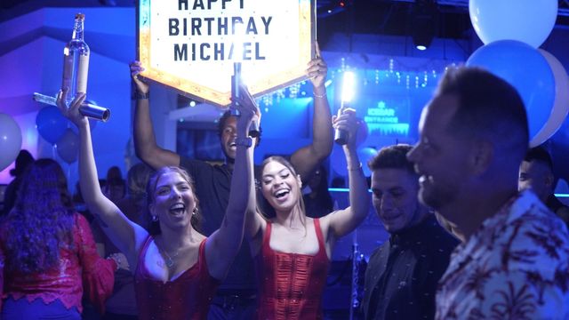 Energetic nightclub birthday celebration with smiling partygoers holding an illuminated birthday sign, bottle service and handheld sparklers under blue club lighting and balloons.