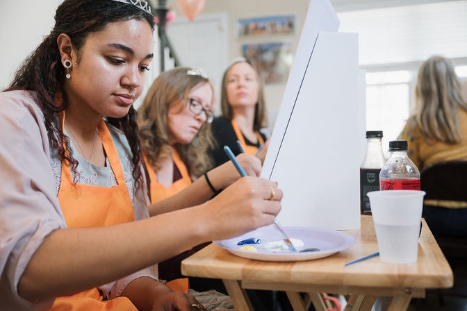 Women in orange aprons at an indoor painting workshop, focused on brushes and a shared palette while working at tabletop easels.