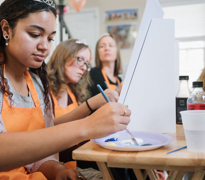 Women in orange aprons at an indoor painting workshop, focused on brushes and a shared palette while working at tabletop easels.